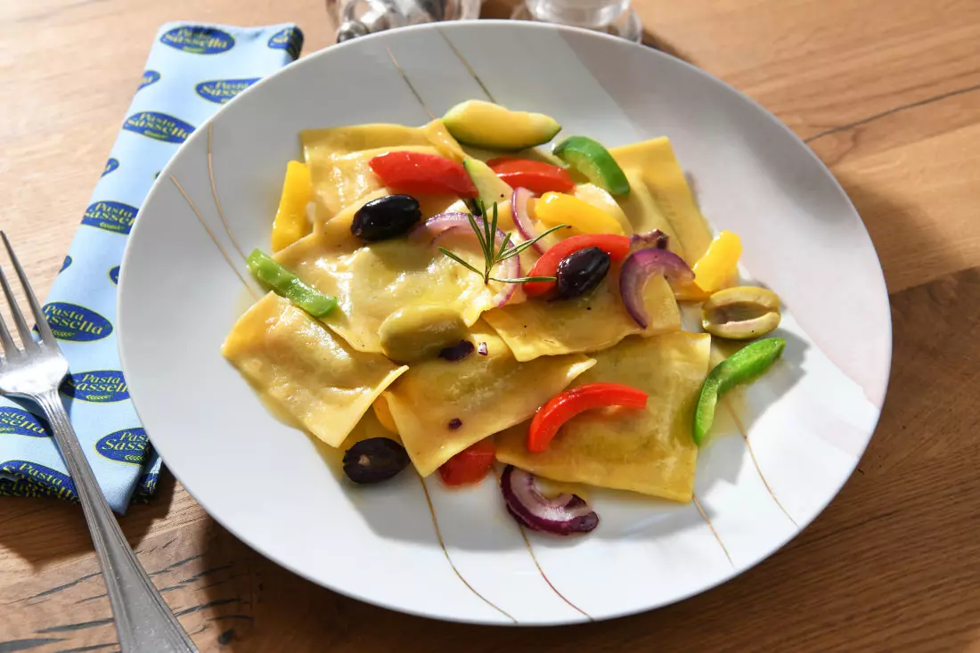 Gefüllte Nudeln (Ravioli) mit Gemüse, Oliven und Zwiebeln auf einem weißen Teller.